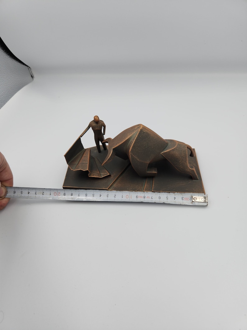 May include: A bronze-colored sculpture of a bullfighter facing a bull. The bull is lying down and the bullfighter is holding a cape. The sculpture is on a rectangular base.