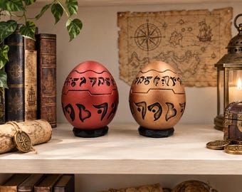 May include: Two egg-shaped containers, one metallic red and one metallic bronze, each featuring horizontal bands of black engraved symbols. Both objects rest on small, black, faceted circular bases and are positioned side by side on a shelf.