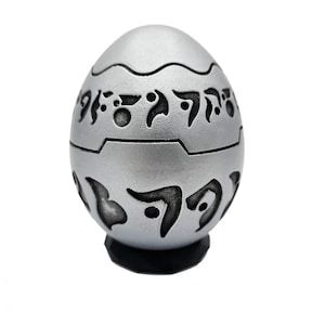 May include: A silver-colored, egg-shaped decorative item with dark etched symbols. The egg has a segmented design with a base and is set against a white background. The symbols appear to be an ancient script.