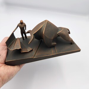 May include: A bronze-colored sculpture of a bullfighter facing a charging bull. The sculpture is made of a textured material and is set on a rectangular base.
