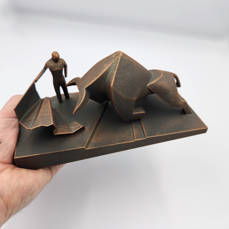 May include: A bronze-colored sculpture of a bullfighter facing a charging bull. The sculpture is made of a textured material and is set on a rectangular base.
