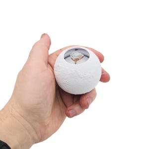 Puede incluir: Una caja de anillos esférica blanca y texturizada con un anillo dentro. El anillo tiene una piedra grande en forma de pera de color azul claro y una banda dorada. La caja se sostiene en una mano sobre un fondo blanco.
