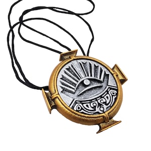 May include: A gold and silver pendant with a black cord. The circular pendant features an eye design with sun rays and decorative patterns. The pendant has a gold border and silver center, creating a striking contrast. The cord is black.