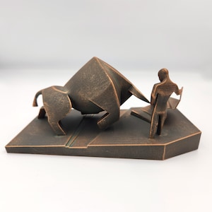 May include: A bronze-colored sculpture of a bull charging towards a person. The bull is stylized and angular, while the person is more realistic. The sculpture is set on a rectangular base.