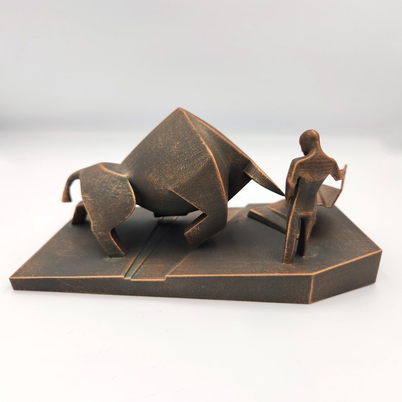 May include: A bronze-colored sculpture of a bull charging towards a person. The bull is stylized and angular, while the person is more realistic. The sculpture is set on a rectangular base.