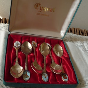 May include: A set of six silver spoons with colorful enamel designs in a green box with a red satin lining. The box has the text "Eygner Souvenirs" on the lid.