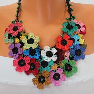 Può includere: Una collana di fiori all'uncinetto colorati con centri neri. La collana è composta da più strati di fiori in vari colori, tra cui rosso, rosa, blu, verde, giallo e bianco.