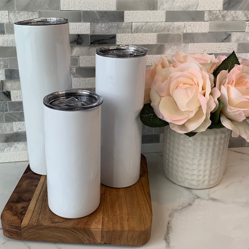 May include: Three white stainless steel tumblers with lids on a wooden surface. The tumblers are different sizes.