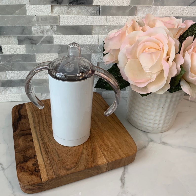 May include: White stainless steel tumbler with a clear plastic lid and two handles. The tumbler is on a wooden cutting board.