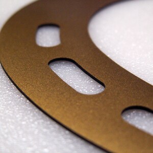 May include: A gold-coloured metal gasket with multiple cutouts. The gasket is on a white textured surface.