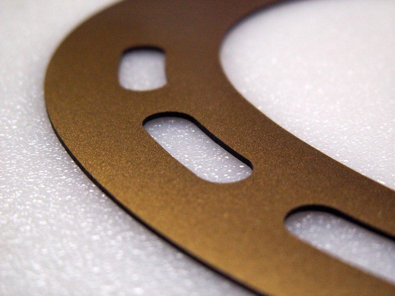 May include: A gold-coloured metal gasket with multiple cutouts. The gasket is on a white textured surface.