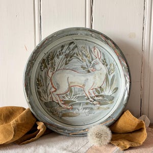 May include: A ceramic plate featuring a hand-painted white rabbit amidst wheat. The plate has a blue-grey rim with red and brown details. It rests on striped fabric, accompanied by a dandelion and a folded brown cloth. The plate's diameter is approximately 25 cm.
