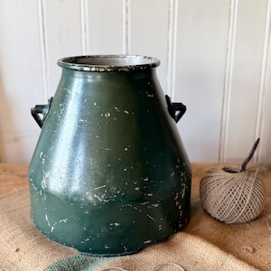 May include: A dark green metal milk churn with a wide opening, two side handles, and a distressed finish showing paint chips. The exterior surface features visible wear and scratches throughout the dark green coating, revealing the metal underneath.