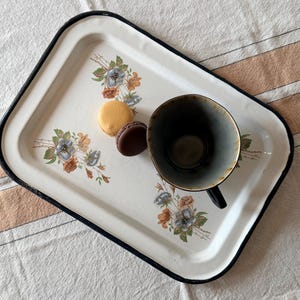 May include: A rectangular white enamel tray with a black rim, decorated with floral patterns in blue and orange. The tray holds one yellow macaron, one dark brown macaron, and a dark grey ceramic mug with a black handle and a dark interior.