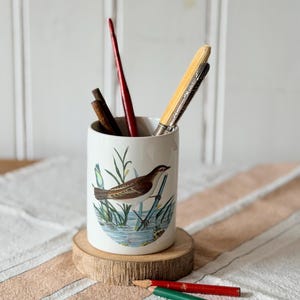 May include: A white ceramic cylindrical pencil holder featuring a detailed illustration of a brown and white bird perched on reeds in water. The holder contains a red pen, a yellow pen, and two dark brown pencils, with a red and green pencil nearby.