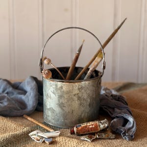 May include: A small, galvanized metal bucket with a wire handle contains paintbrushes. A wooden object with a floral design is inside. Paint tubes and broken ceramic pieces are on a burlap surface, evoking an artist's studio.