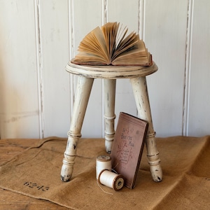 May include: A small, three-legged wooden stool with a distressed white finish. An open book with yellowed pages rests on the seat. Below, a brown book titled The Babe in the Mill leans against the legs next to two spools of white thread.