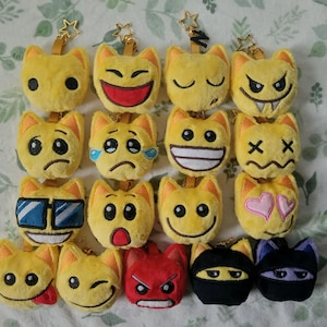 May include: A selection of yellow cat-shaped emoji keyrings with diverse facial expressions, such as smiling, crying, and angry. Some keyrings feature sunglasses or hearts. Each keyring has a gold star-shaped clasp.