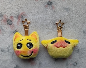 Può includere: Due portachiavi in ​​peluche gialli con facce ricamate. Uno ha una faccia sorridente con guance rosee, l'altro un'espressione sorpresa. Entrambi hanno un attacco portachiavi a forma di stella dorata e un cinturino in pelle marrone.