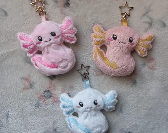 Puede incluir: Tres llaveros de peluche de axolotl. Uno es rosa con purpurina dorada, otro es rosa con purpurina blanca y el último es blanco con purpurina azul. Cada axolotl tiene un amuleto de estrella dorada unido a la parte superior.