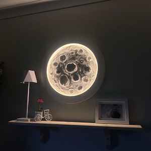 Può includere: Un'opera d'arte da parete a forma di luna rotonda e illuminata con un design dettagliato dei crateri. Una piccola fioriera a forma di bicicletta bianca con fiori rossi è appoggiata su una mensola, insieme a una foto con cornice bianca e una lampada moderna.
