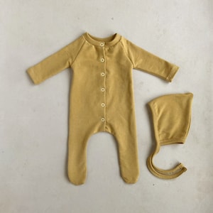 Könnte beinhalten: Ein senfgelber Baby-Strampler mit Knopfleiste und passender Mütze. Der Strampler hat lange Ärmel und Fußspitzen.