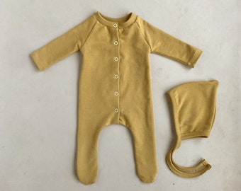 May include: A mustard yellow baby onesie with a button closure and matching hat. The onesie has long sleeves and footed legs.