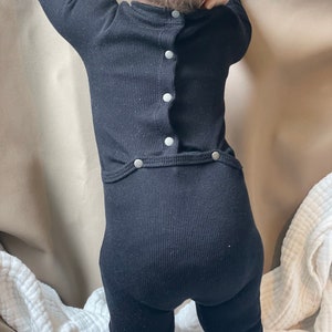 Könnte beinhalten: Schwarzer gerippter Baby-Strampler mit einer Reihe silberner Knöpfe auf dem Rücken.