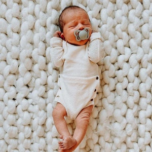 May include: A newborn baby wearing a white onesie and a gray pacifier, lying on a white fluffy blanket.