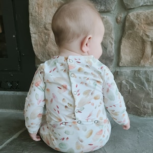 May include: A white long-sleeved baby onesie with a watercolor print of leaves and flowers in shades of pink, orange, and green. The onesie has a button closure on the back.