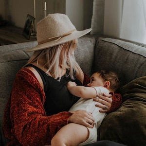 May include: A woman wearing a brown hat and a red cardigan is holding a baby in her arms. The baby is wearing a white onesie and is looking at the woman. They are sitting on a grey sofa.