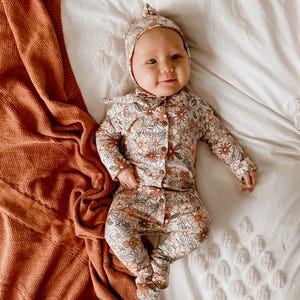 Op de afbeelding: Een baby draagt een wit en bruin bloemenprint onesie en bijpassende muts. De onesie heeft een knoopsluiting en de muts heeft een geknoopte bovenkant. De baby ligt op een wit laken met een bruin gebreid deken aan de linkerkant.