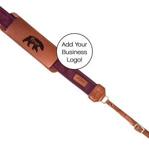 Può includere: Tracolla per fotocamera color bordeaux con una toppa in pelle marrone che dice "Mama" e una silhouette di un orso. La tracolla ha un anello in metallo dorato e una fibbia dorata.