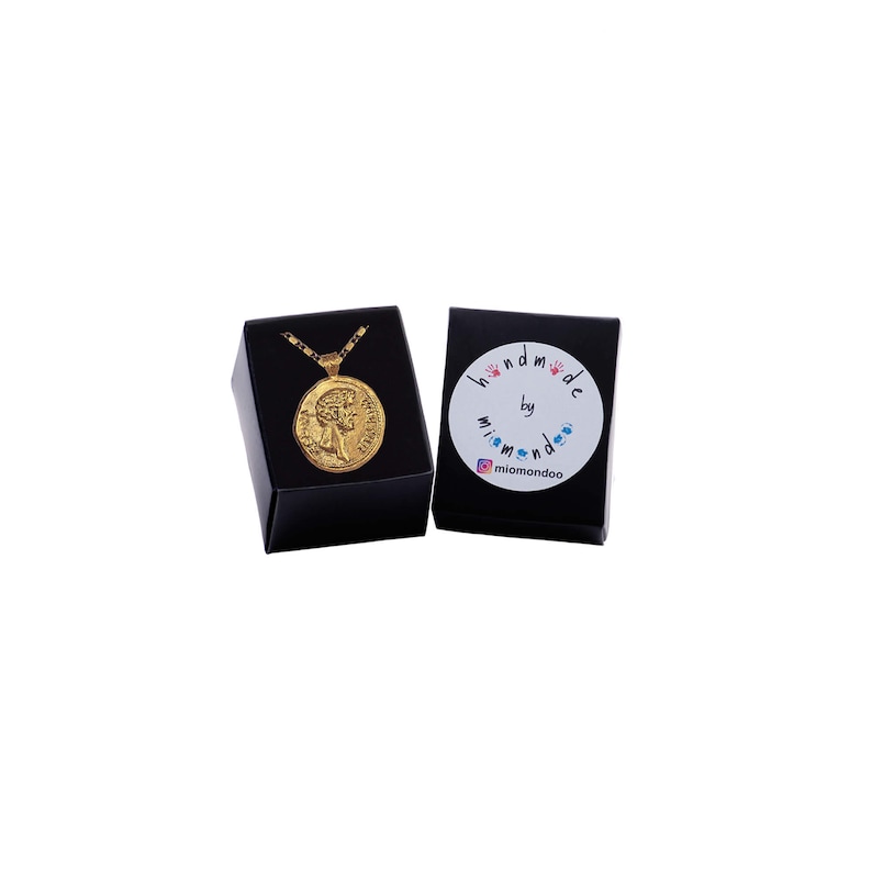 May include: Gold necklace with a round pendant featuring a profile of a woman's face. The necklace is in a black box with a white label that says "handmade by mimomondoo".