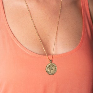 May include: A gold chain necklace with a gold coin pendant featuring a profile of a person's face. The necklace is worn by a person with a peach coloured tank top.