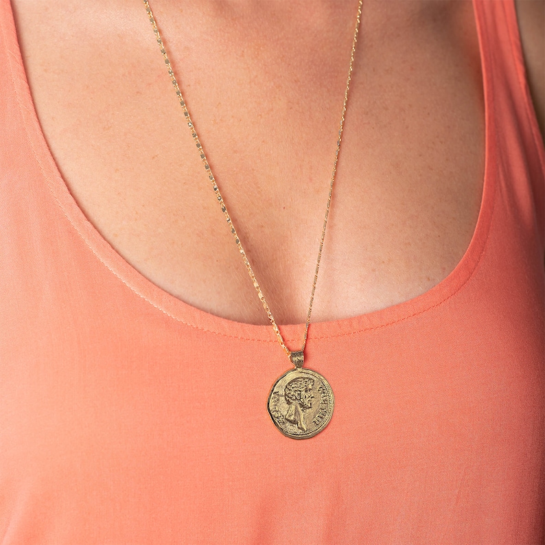 May include: A gold chain necklace with a gold coin pendant featuring a profile of a person's face. The necklace is worn by a person with a peach coloured tank top.