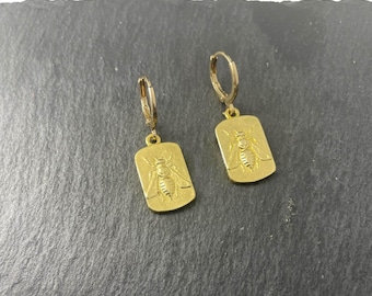 May include: A pair of gold-toned hoop earrings featuring rectangular pendants. Each pendant displays a detailed embossed bee design on the front surface. The earrings are constructed with a hinged closure mechanism for secure attachment to the earlobe.