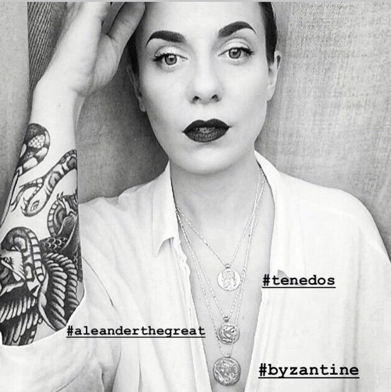 May include: A black and white photograph of a person wearing a white shirt with several silver necklaces. The necklaces feature coin-shaped pendants with intricate designs. The person has a black and white tattoo on their arm, featuring a snake and a bird. The image includes the hashtags #aleanderthegreat, #tenedos, and #byzantine.