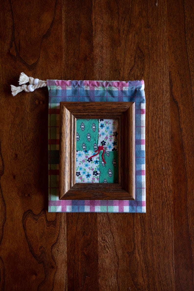 May include: A small framed picture with a patchwork design of green and floral fabric. The frame is dark brown wood and the picture is sitting on a pink and blue plaid fabric.