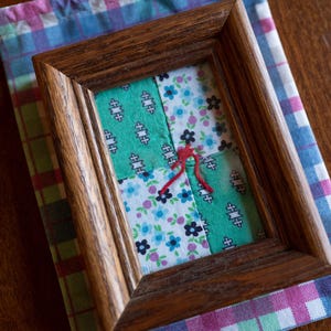 May include: A framed patchwork quilt with a red string in the center. The quilt is made of four squares of fabric, two green, one blue with white flowers, and one white with blue flowers. The frame is dark brown wood.