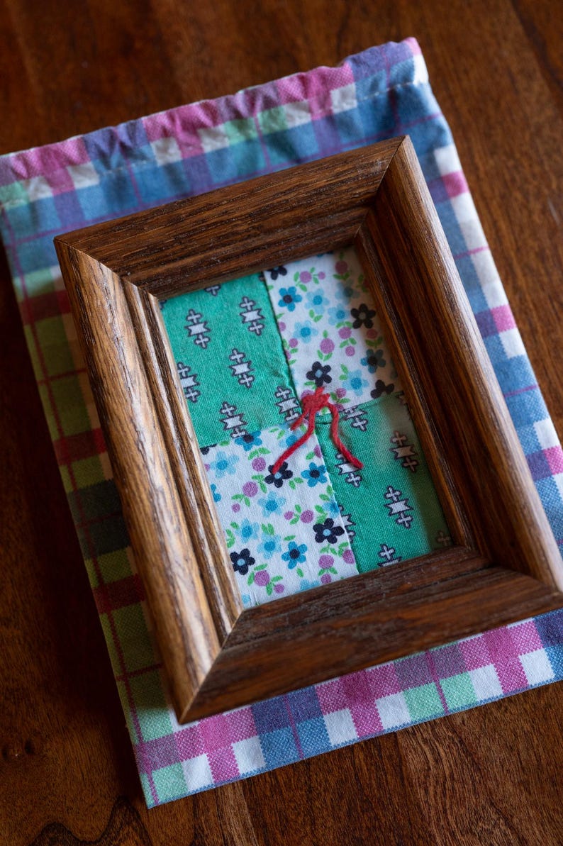 May include: A framed patchwork quilt with a red string in the center. The quilt is made of four squares of fabric, two green, one blue with white flowers, and one white with blue flowers. The frame is dark brown wood.