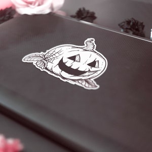 May include: A black and white sticker of a jack-o'-lantern with a spider web design. The sticker is on a dark gray laptop. The image is decorated with pink and black flowers and a lit candle.