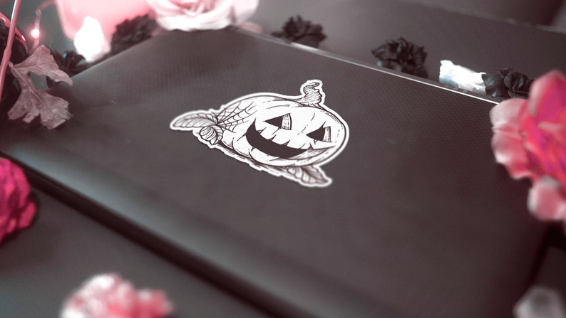 May include: A black and white sticker of a jack-o'-lantern with a spider web design. The sticker is on a dark gray laptop. The image is decorated with pink and black flowers and a lit candle.