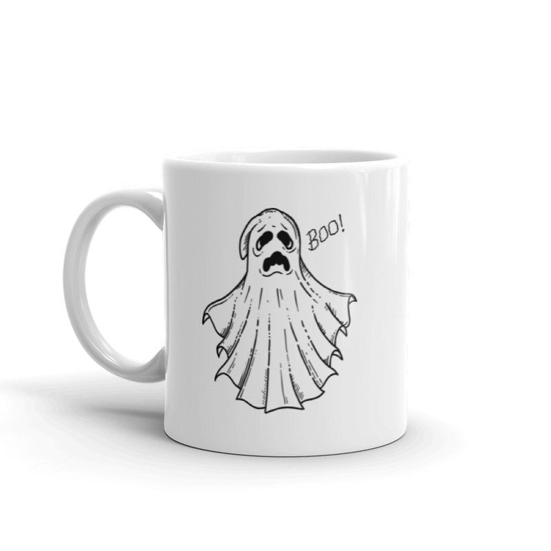 May include: White ceramic mug featuring a black and white illustration of a cartoon ghost with a sad expression. The word "Boo!" is written next to the ghost. The mug has a curved handle.