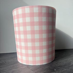 May include: A cylindrical container featuring a pink and white gingham pattern. The pattern consists of evenly spaced squares in pink and white. The container is likely made of paper or cardboard, and is a decorative item.