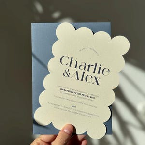 May include: A wedding invitation featuring the names "Charlie & Alex" in elegant typography. The invitation has a scalloped edge and is cream-colored, set against a blue background. The text includes the wedding date, time, and location.