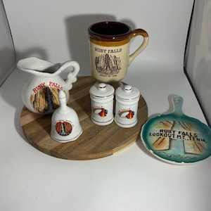 Könnte beinhalten: Eine Sammlung von Ruby Falls Souvenirs, darunter eine Tasse, ein Sahnekännchen, eine Glocke, Salz- und Pfefferstreuer und ein kleiner Teller. Die Artikel sind weiß, beige und türkis, mit Illustrationen von Ruby Falls und Lookout Mountain, Tennessee.