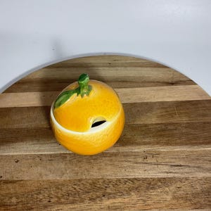 Puede incluir: Un tarro de cerámica con forma de naranja con tapa y una pequeña abertura. El tarro es de color naranja brillante con una hoja y un tallo verdes en la parte superior. El tarro está sobre una superficie de madera.