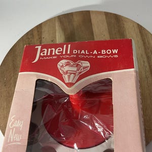 以下が含まれることがあります： 赤とピンクの厚紙箱に入ったJanell Dial-A-Bowクラフトツール。箱にはJanell DIAL-A-BOW MAKE YOUR OWN BOWSおよびEasy Newの文字が記載されています。透明なプラスチック窓から、白い縁取りのある赤い円形のプラスチック製ベースが見えます。