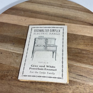 May include: A vintage booklet for a Buckwalter-Simplex electric range. The cover features a black and white illustration of the appliance. Text on the cover reads "Gray and White Porcelain Enamel For the Large Family". The booklet is on a wooden surface.